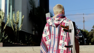 Organic serape and wrap with hood worn by surfer holding surfboard on beach next to cactus with blond hair in buzz cut